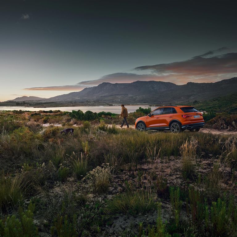 An orange Audi Q3 parked on a dirt path near a lake. A man is walking away from the car with his dog.